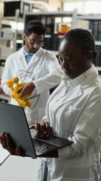 Vertical video of young adult African American female scientist typing on laptop while colleague works on robotic device. Clip is ideal for engineering education and technology marketing
