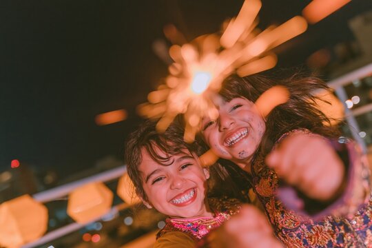 Two South Asian children laughing while holding glowing sparklers on a balcony, featuring embroidered clothing and amber lanterns for Diwali and family celebrations