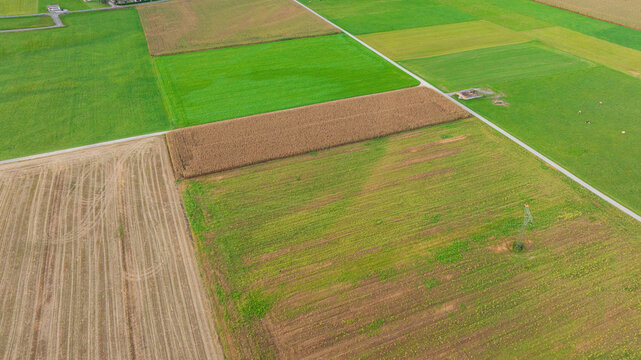 High angle aerial shot displays scenic rural landscape of Lind im Drautal village. Winding Drava river, agricultural fields, main road, and railway line cross lush green valley surrounded by forested 