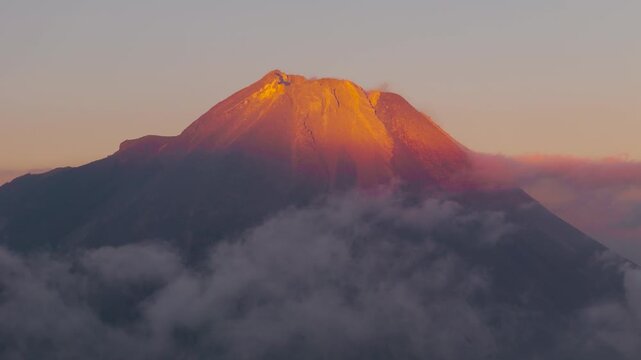 Aerial view of Mount Merapi glowing in golden sunrise light with clouds drifting around the volcanic slopes in Dukun, Central Java, Indonesia.