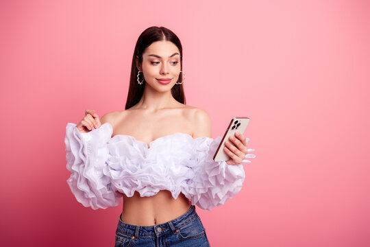 Young woman in ruffled white crop top holding smartphone against pink background with a playful confident expression