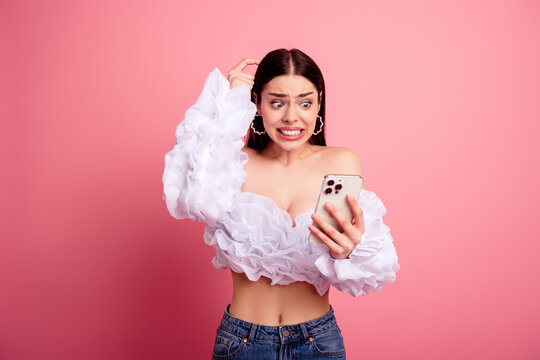 Young woman in ruffled white crop top and jeans looking shocked and confused at her smartphone while scratching her head against a pink background