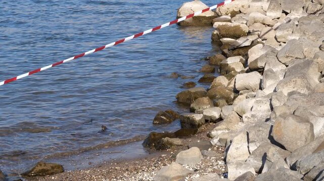 A close-up, diagonal shot of a red and white warning barrier tape stretched across a scenic shoreline filled with large grey rocks and a flowing blue river. The restriction tape creates a sense of cau