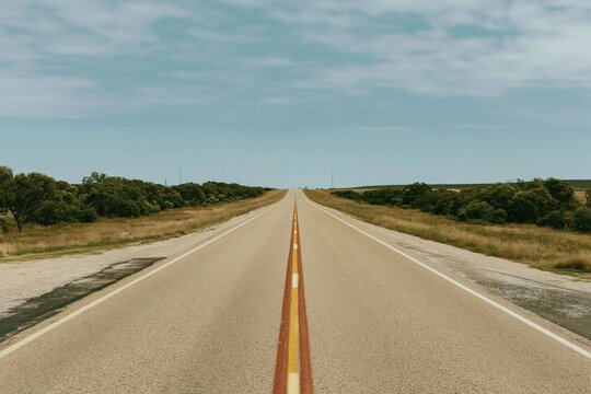 Empty asphalt highway with worn orange markings leading toward a distant skyline under a muted blue sky, for infrastructure and travel banners