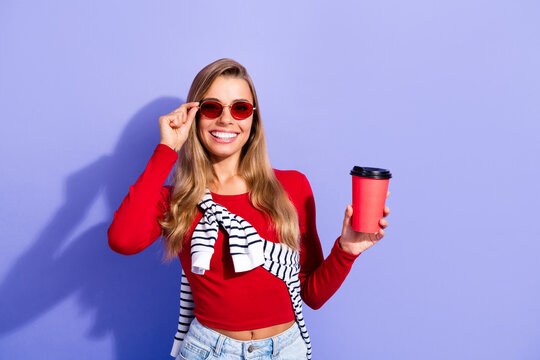 Smiling young woman in red top holding takeaway coffee cup and wearing sunglasses for casual fashion lifestyle portrait against a purple background