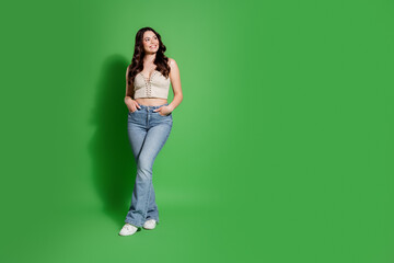 Smiling young woman in casual crop top and jeans posing against a bright green studio background,...
