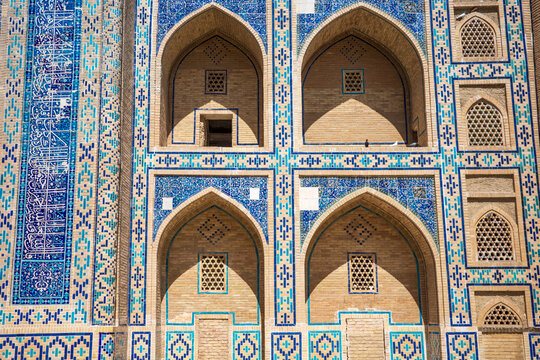 Detailed facade of a historic madrasa in Bukhara, Uzbekistan, featuring symmetrical pointed arches, turquoise and blue mosaic tilework, geometric brick patterns, and decorative Islamic calligraphy