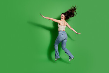 Young woman jumping and smiling in casual jeans against a bright green studio background, joyful...