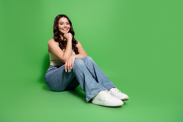 Smiling young woman in casual outfit sitting on green background with jeans and white sneakers,...