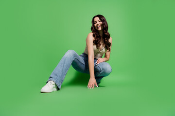 Young woman in casual jeans and white sneakers smiling while crouching on a vibrant green studio...
