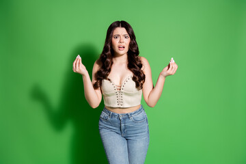 Confused young woman shrugging and gesturing in front of a green background expressing surprise and...