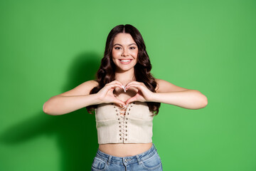 Smiling young woman making heart gesture with hands in front of green background, casual portrait...