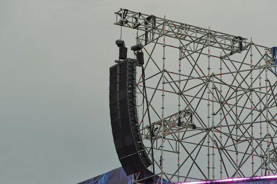 Professional line array speakers hanging from metal stage truss structure against overcast sky.