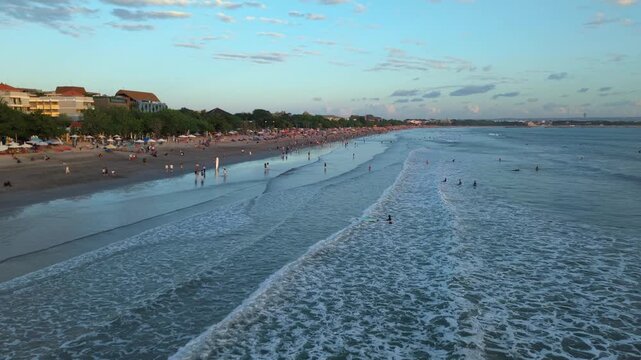 Aerial view of Kuta Beach with gentle waves rolling onto the sandy shore, people walking along the coastline, and buildings lining the beach in Kuta, Bali, Indonesia.