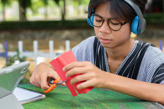 A young man with blue headphones and glasses cutting red paper with scissors outdoors, focused on an art craft project with a tablet nearby.