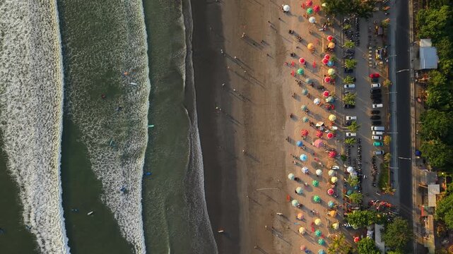Aerial view of Kuta Beach with rows of colorful umbrellas on the sand and surf waves rolling onto the shore in Kuta, Bali, Indonesia.