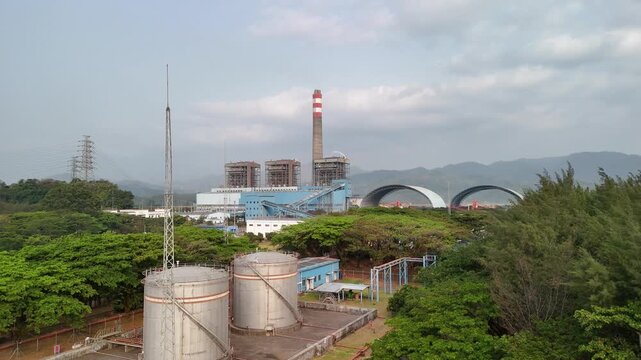 Aerial view of a power plant with large storage tanks, a tall chimney, and surrounding lush greenery