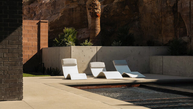 Modern white lounge chairs positioned beside a tranquil swimming pool against a rustic desert rock wall and concrete barrier