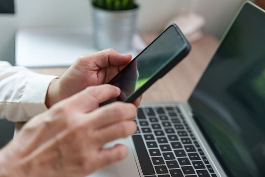 Close-up of a person using a smartphone beside an open laptop at a desk, showing mobile communication, online browsing, digital technology, remote working, multitasking and modern lifestyle.
