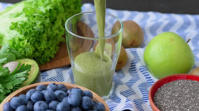 Green smoothie pours into glass on kitchen counter among healthy organic ingredients: lettuce, blueberries, kiwi, chia seeds, green apple. Concept: healthy breakfast, homemade, healthy lifestyle