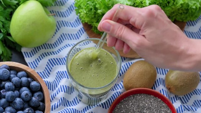 Woman hand mixes green smoothie in glass using straw, with blueberries, kiwi, apple, leafy greens and chia nearby, presenting fresh nutrition, balanced eating and healthy beverage preparation.