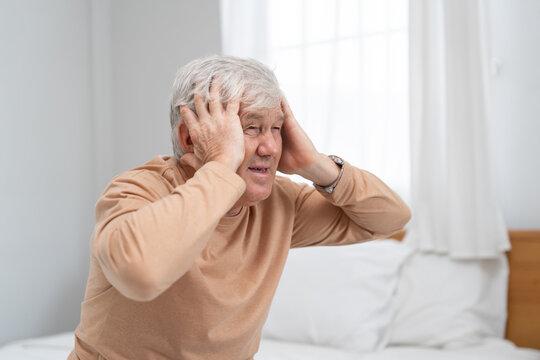 Elderly man sitting on bed holding head suffering painful headache migraine, senior male feeling stressed memory loss dizziness healthcare medical concept