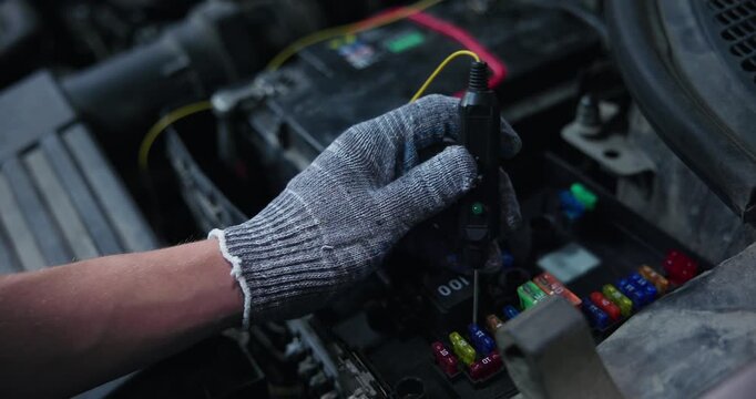 Handheld tracking shot of gloved hand probing car fuse box under open hood with voltage tester and red indicator light, close steady movement.