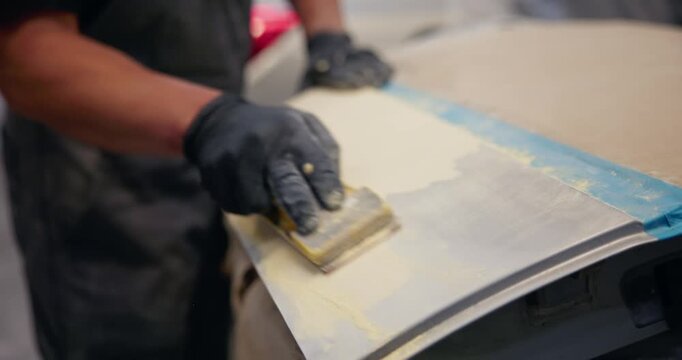 Handheld sanding of car door panel in auto body workshop with protective gloves.