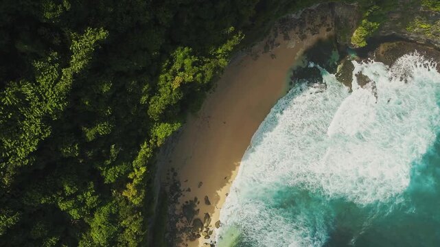 Aerial view of Pantai Nunggalan with turquoise waves crashing onto the sandy shore, bordered by lush tropical forest cliffs in Kecamatan Kuta Selatan, Bali, Indonesia.