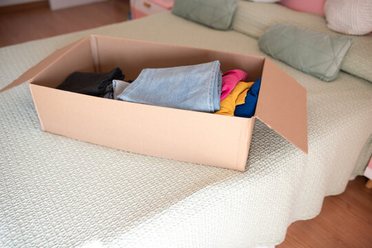 Individual placing folded clothes into a cardboard box on a bed with a light green cover