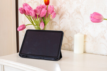 A tablet in a black case stands beside a candle and a vase of pink and orange tulips on a white table, a soft cozy scene © Дмитрий Швец