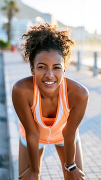 Young Black woman in colorful running gear pauses on beach promenade, smiling while catching breath with coastal scenery in background