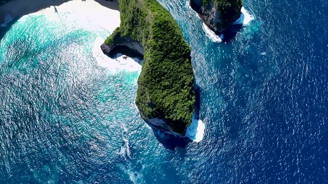 Aerial view of the iconic T-Rex shaped cliff of Kelingking Beach surrounded by deep blue ocean waves crashing against the rocky coastline in Nusa Penida, Bali, Indonesia.