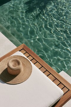 Woven straw hat placed on a teak sun lounger beside a turquoise swimming pool in a Tropical Modernism villa setting