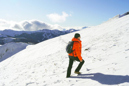 Hiker in bright orange jacket trekking up snowy mountain slope at Kolasin 1600 ski resort. Illustrates winter summit ascent, off-piste recreation, and alpine wilderness exploration during snow season.