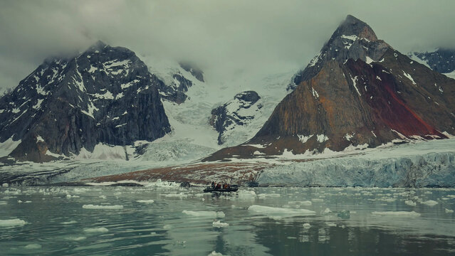 A small zodiac boat with tourists exploring an icy bay filled with floating ice floes beneath towering mountains