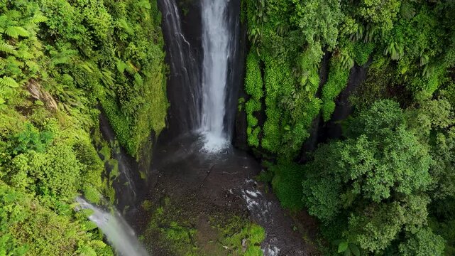 Aerial view of Curug Ciarjuna waterfall cascading down a lush green cliffside surrounded by dense tropical vegetation in Garut, West Java, Indonesia.