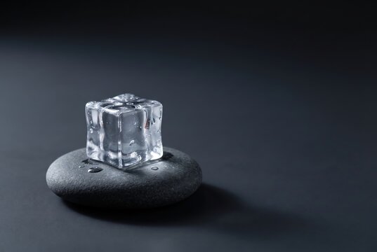 A clear ice cube rests on a smooth, dark stone against a dark background, highlighting textures and reflections in minimalistic, tranquil composition.