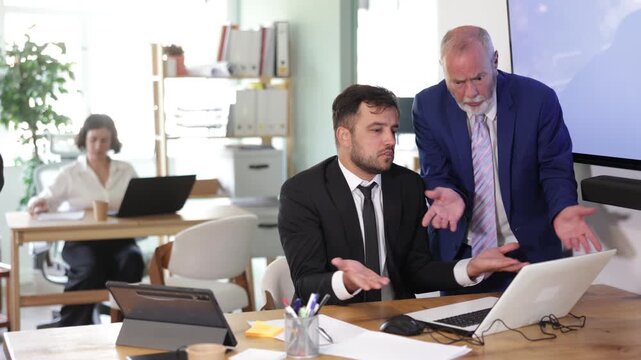 Tense head of the office is angry and scolds at the male employee, he explains to him the work tasks and deadlines. Elderly man standing in office and swearing at colleague near laptop . High quality
