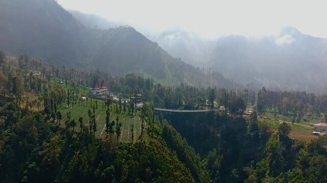 Aerial view of the Jembatan Kaca glass bridge spanning a lush mountain valley with dense trees and misty slopes in Kecamatan Sukapura, Jawa Timur, Indonesia.