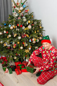 A 53 year old man sits near a decorated Christmas tree on Christmas Day. He is opening a gift while wearing a festive outfit and a holiday hat. Family and joy fill the space around him