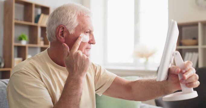 Senior man applying skincare patches at home. Elderly male looks in mirror while grooming, targeting under eyes and reducing puffiness, wrinkles, and fatigue. Male beauty and wellness concept.
