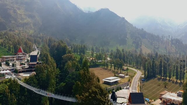 Aerial view of Jembatan Kaca glass bridge spanning a lush valley with surrounding forest and mountain landscape in Kecamatan Sukapura, Jawa Timur, Indonesia.