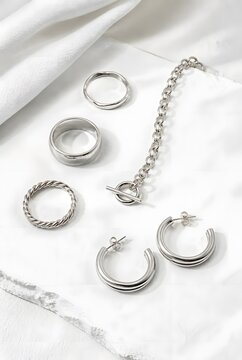 Flat Lay of Silver Rings Chain Bracelet and Hoop Earrings on White Textured Fabric Background