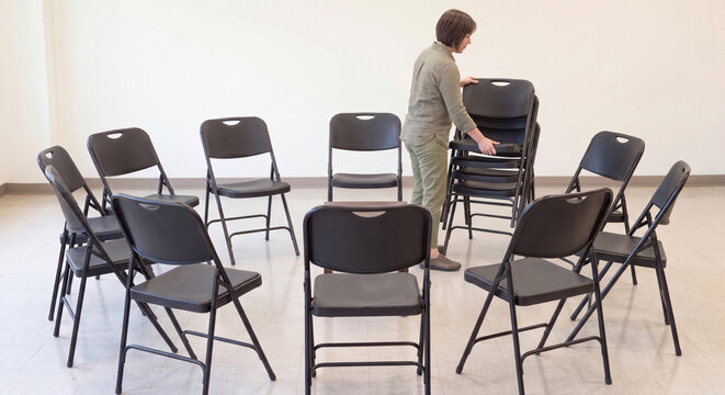 Asian woman arranges and stacks black folding chairs into a circle layout inside a bright and minimalist empty workshop studio