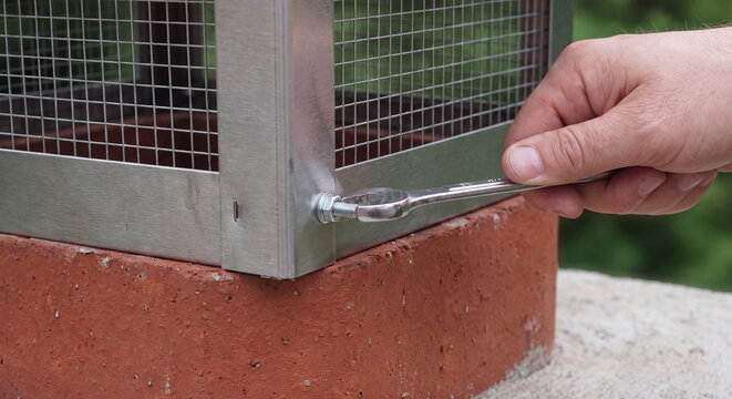 A technician uses a spanner wrench to securely fasten a stainless steel spark arrestor cap onto the red brick chimney of a residential rooftop