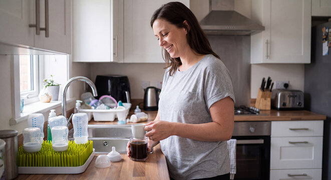 A focused mother prepares baby formula milk in a kitchen bottle surrounded by feeding equipment and drying rack on the wooden counter