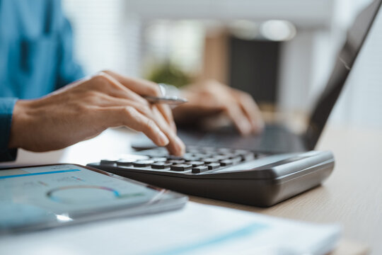 Two colleagues use a calculator and laptop for finance planning, combining budgeting, accounting, expense and cost analysis in a report that helps their business compare revenue and finance goals.