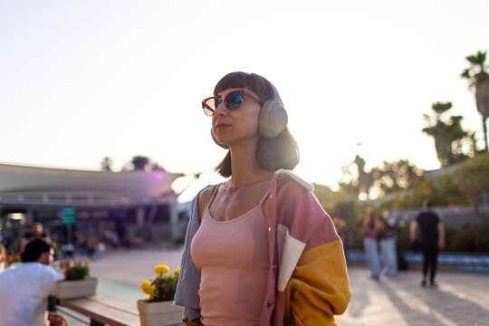 A woman wearing headphones and sunglasses enjoys her favorite song on a sunlit street. The woman smiles as she listens to the music and shows off her rings and bracelets.