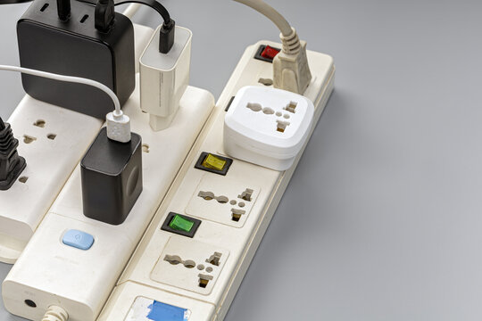 Close-up of an overloaded white electrical power strip with multiple black and white USB chargers, adapters, and universal plug sockets, illustrating electrical safety hazards and overload risk